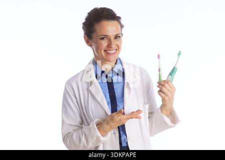 A female dentist in a lab coat and tie gestures while holding toothbrushes, providing educational advice on proper dental hygiene techniques. Stock Photo