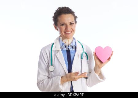 A cheerful doctor in a lab coat holds a pink heart and gestures toward it, promoting heart disease prevention and compassionate cardiovascular healthc Stock Photo