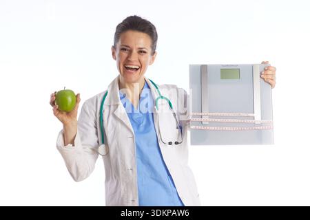 A smiling doctor in medical attire holds a digital scale and a green apple, symbolizing a positive and professional approach to weight loss and health Stock Photo