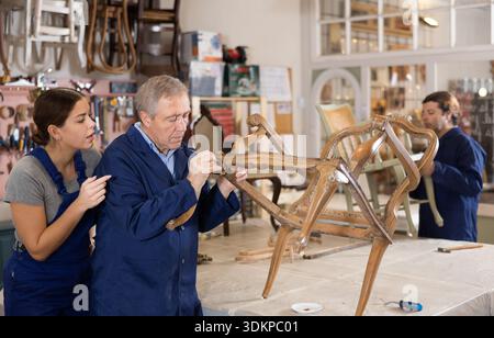 Senior male furniture maker and female apprentice working on vintage commode in workshop Stock Photo