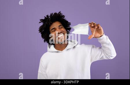 Happy black guy taking off medical mask, feeling free on violet studio background Stock Photo