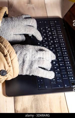 Vertical close up side view of warm gloved hands typing on a laptop during winter blackout, showing remote work and home office lifestyle in Ukraine. Stock Photo