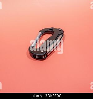 A black multi-tool carabiner with silver components lies diagonally on a coral pink background, showcasing its utility features. Stock Photo