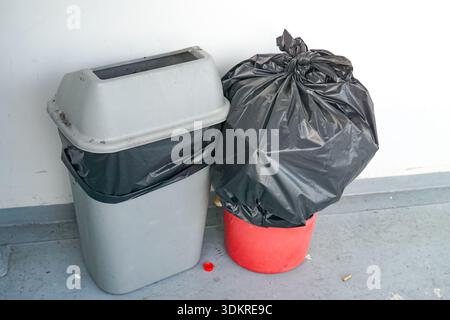Two trash bins side by side on a gray floor against a white wall, one properly used and one overflowing, highlighting waste management and cleanliness Stock Photo