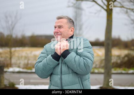 Mature man standing outdoors in winter, holding his jacket collar and looking into the distance with a thoughtful and slightly concerned expression ag Stock Photo