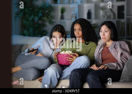 Three diverse amazed friends watching tv in the night sitting on a couch at home Stock Photo