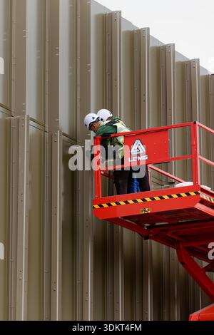 Two contractors with ppe on scissor lift fix aluminium fins to vertical ...