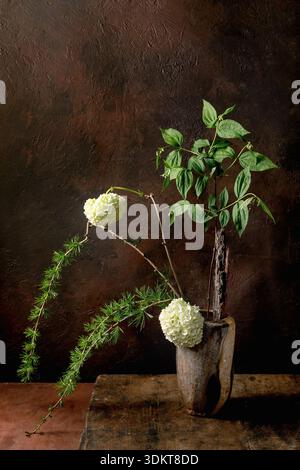 Artistic still life with ikebana white hydrangea flowers green foliage and pine branches in vintage wooden vase against dark textured background. Eleg Stock Photo