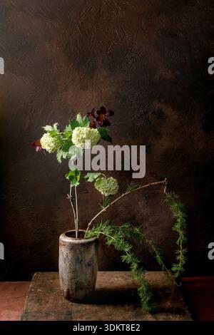 Artistic still life with ikebana white hydrangea flowers and pine branches in rustic ceramic vase against textured dark brown background. Natural comp Stock Photo