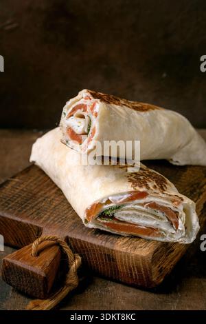 Delicious salmon lavash wrap with cream cheese fresh greens and vegetables served on rustic wooden cutting board. Homemade appetizer with smoked salmo Stock Photo