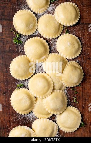 Homemade ravioli pasta with flour on rustic wooden cutting board top view. Fresh Italian pasta ready for cooking with herbs decoration. Stock Photo