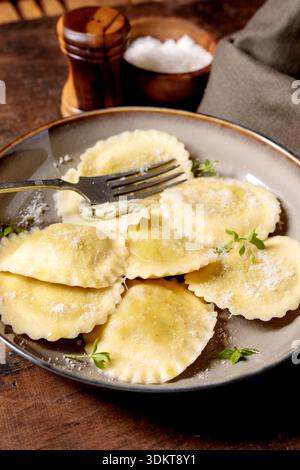 Fresh homemade ravioli pasta filled with cheese served on ceramic plate with fork garnished with herbs and parmesan cheese on rustic wooden table Stock Photo