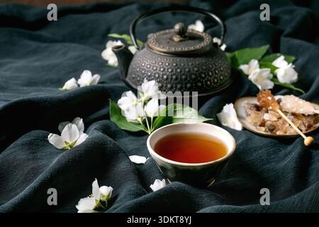 Traditional tea ceremony setup with cast iron teapot cup of tea brown sugar jasmine flowers on dark fabric background Stock Photo