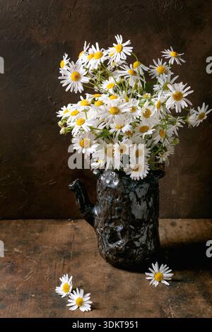 Fresh white daisies arranged in dark ceramic vase on wooden surface with scattered petals rustic home decor style Stock Photo