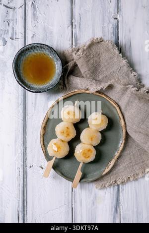 Top view Japanese dango dumplings on ceramic plate with tea bowl wooden sticks linen napkin on white wooden background. Traditional Asian dessert Stock Photo