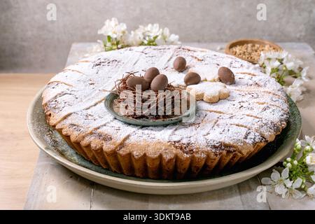 Homemade Easter tart Pastiera Napolitano dusted with powdered sugar decorated with chocolate nest and eggs surrounded by white spring flowers on woode Stock Photo