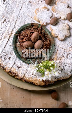 Traditional Easter dessert with chocolate eggs in nest on powdered traditional pie Pastiera Napolitano and white spring flowers on rustic background. Stock Photo