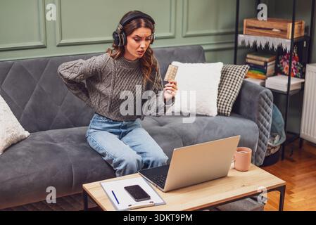 Young woman is sitting on bed in living room and having online doctor's appointment using laptop. Young woman taking medication while working on lapto Stock Photo