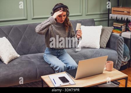 Young woman is sitting on bed in living room and having online doctor's appointment using laptop. Young woman taking medication while working on lapto Stock Photo