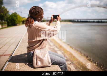 Young cheerful black woman is sitting by the river in the city and taking photos. Stock Photo