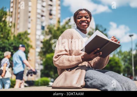 Cheerful African American woman relaxing in the city park, reading a book. Stock Photo