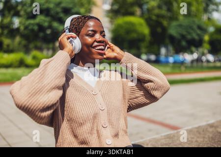 Portrait of happy and joyful African American woman who is listening music on wireless headphones on sunny day. Stock Photo