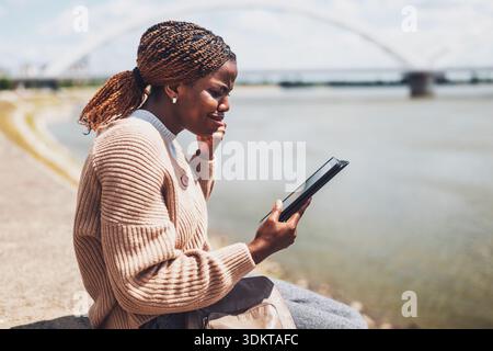 Young worried black woman is using digital tablet. She is sitting by the river in the city. Stock Photo