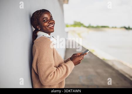 Outdoor portrait of happy young black woman who is messaging on smartphone. Stock Photo