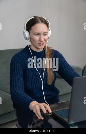 Young woman wearing headphones, engaging in a video call while working from her home office. Focused on her laptop, she talks and navigates with a com Stock Photo