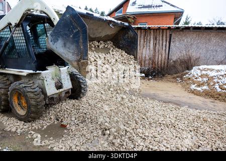 A small construction loader dumps crushed stones on a dirt surface near a red brick house with a snow covered roof and wooden fence in a winter settin Stock Photo