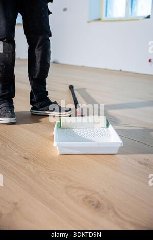 A person stands near a wooden floor being treated, with a paint roller in a tray. Natural light enters through a window, highlighting unfinished walls Stock Photo