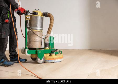 A worker in black attire operates a floor polishing machine on a light wooden surface, with visible tools, red gloves, and blue protective footwear. Stock Photo