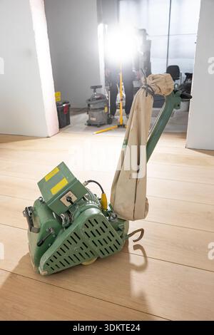 A green floor polishing machine with a beige dust bag on a light wooden floor. Construction tools and a work light are visible in the background. Stock Photo
