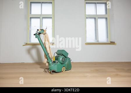 A green floor polishing machine on a light wooden floor in a white walled room. Two large windows with protective plastic suggest ongoing maintenance. Stock Photo