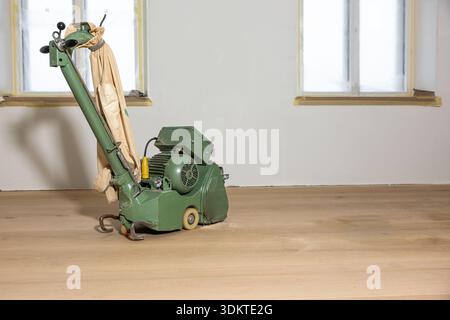A green floor polishing machine with a beige dust bag stands on a smooth wooden floor. Two taped windows and plain light walls suggest ongoing mainten Stock Photo