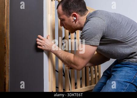 A man in a gray t shirt and blue jeans adjusts a wooden crib frame indoors. The setting includes a gray wall and wooden panel, emphasizing a home envi Stock Photo