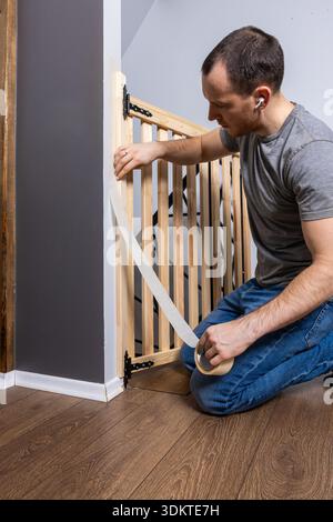 A man kneels on a wooden floor, securing a baby gate with tape. The gate has black hinges and is attached to a gray painted wall near a staircase. Stock Photo