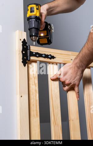 Close up of hands operating a yellow and black cordless drill to attach a black hinge to a light wooden gate with vertical slats indoors. Stock Photo