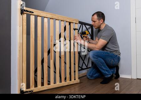 A man kneels while using a yellow and black power drill to install a wooden safety gate with black metal hinges near a staircase with geometric railin Stock Photo