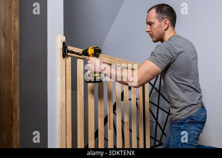 A man uses a yellow and black power drill to secure a light wood baby gate with black hinges to a white wall near a staircase with metal railings. Stock Photo