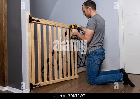 A man kneels on a wooden floor using a yellow and black power drill to install a wooden safety gate with black metal hinges in a modern interior. Stock Photo