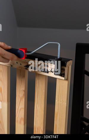A hand uses a red and black paint roller to apply black paint to a wooden slatted structure, showing a contrast between painted and unpainted wood. Stock Photo