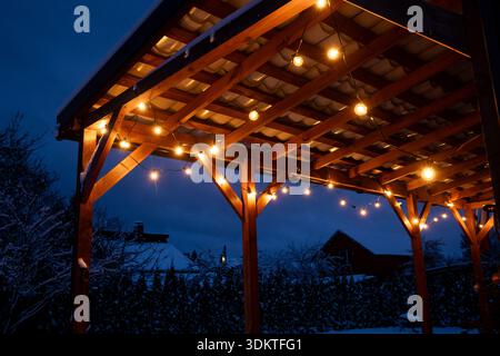 A wooden terrace illuminated by warm LED string lights, surrounded by snow covered trees and rooftops under a dark blue evening sky. Stock Photo