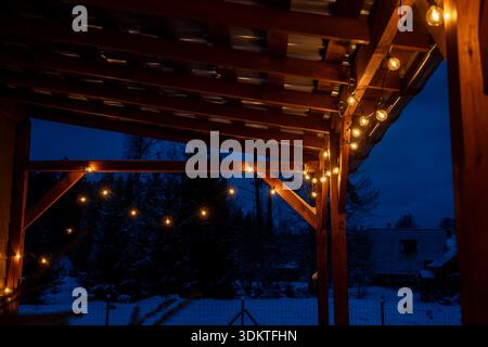 A wooden terrace illuminated by warm LED string lights, surrounded by snow covered ground and trees, with a house and fence in the background. Stock Photo