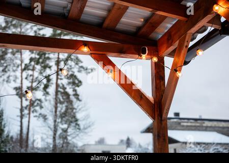 A wooden terrace with string lights glowing warmly, corrugated metal roof, tall trees, snow covered ground, and a modern flat roof building in view. Stock Photo