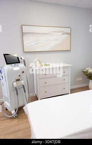 Clean interior featuring a white bed, medical device with touchscreen, white dresser with gold knobs, abstract artwork, and wooden flooring. Stock Photo