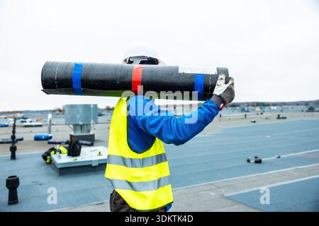 A worker in a white hard hat and yellow vest carries rolled roofing material on a flat rooftop with tools, ventilation units, and a distant horizon. Stock Photo