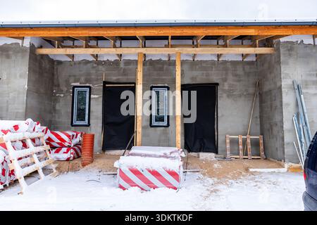 A snow covered construction site featuring a concrete structure with narrow windows, a black covered doorway, wooden beams, and stacked materials. Stock Photo