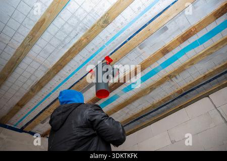 A worker in a blue hood and black jacket installs a red and gray ventilation pipe on a ceiling with exposed wooden beams and a vapor barrier. Stock Photo
