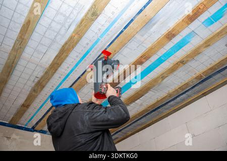 A worker in a blue hood and black jacket uses a power drill to install a black and red ventilation system on a ceiling with wooden beams and insulatio Stock Photo